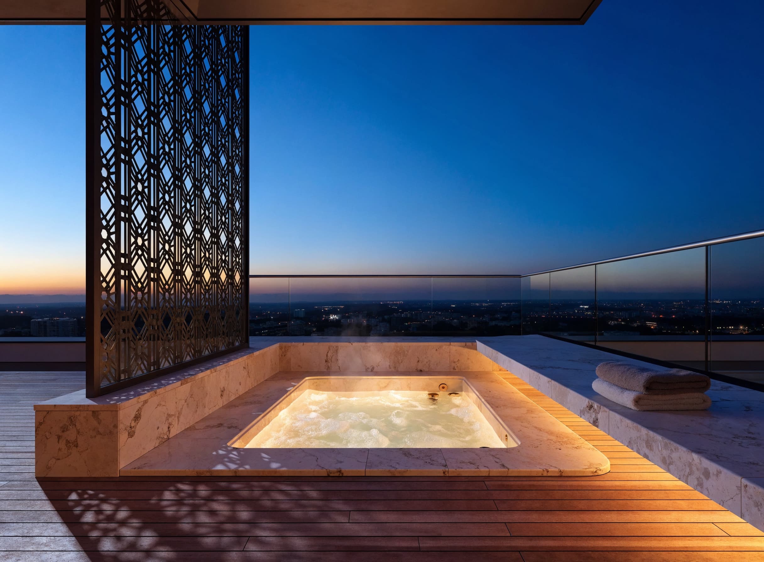 A rooftop jacuzzi at dusk, set in travertine beside a star lattice screen, with the city beyond