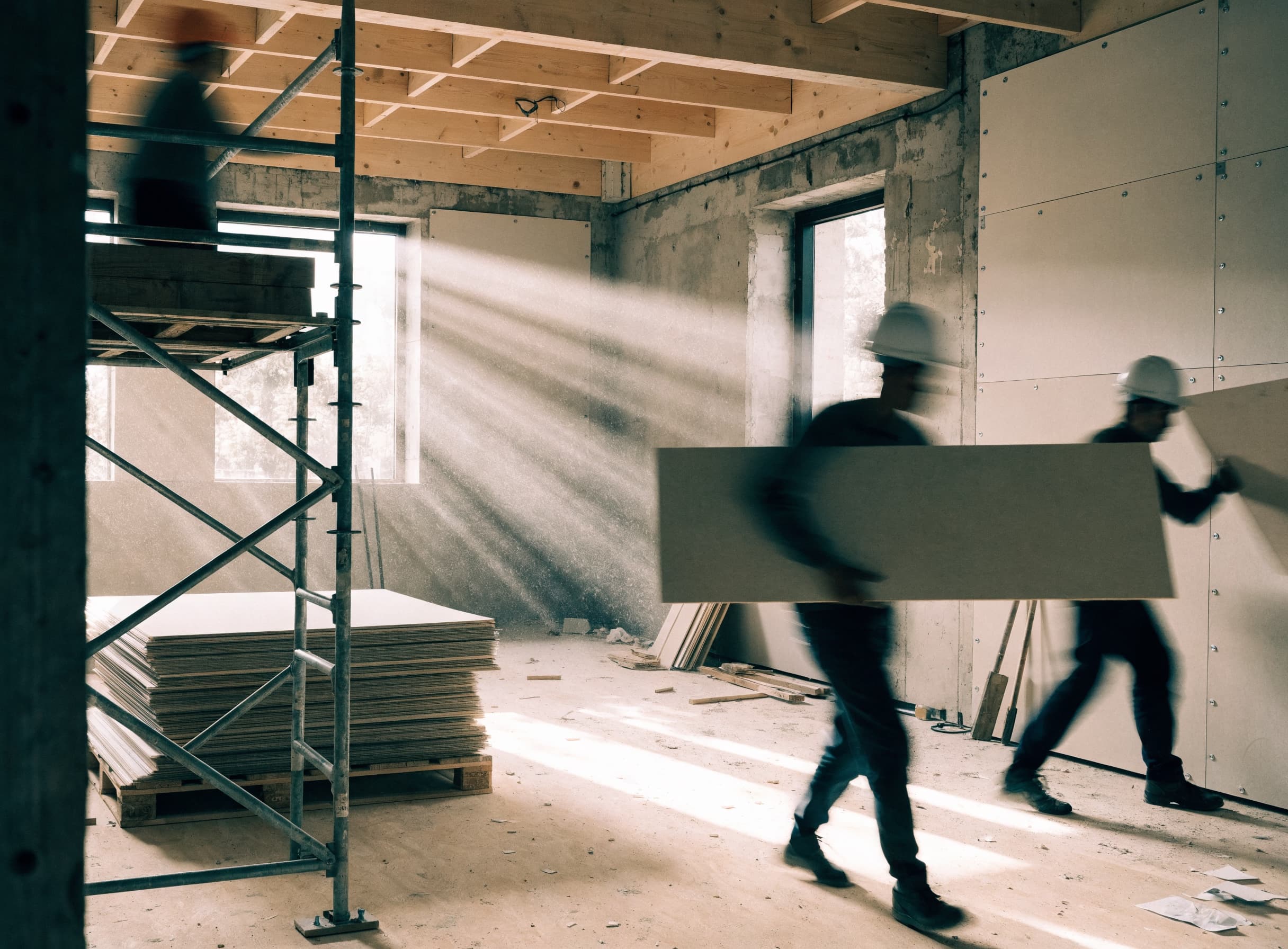 A fit-out underway, workers carrying and fixing panel board, daylight raking through the openings